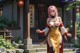 A woman in an elaborate outfit stands outside a traditional building with Chinese lanterns and greenery.